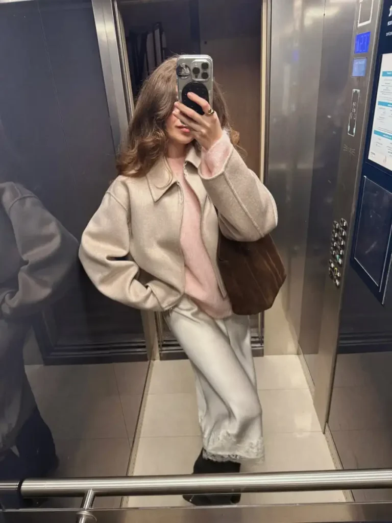 Beige cropped jacket layered over a blush pink sweater and ivory satin skirt with lace trim and a brown suede bag.