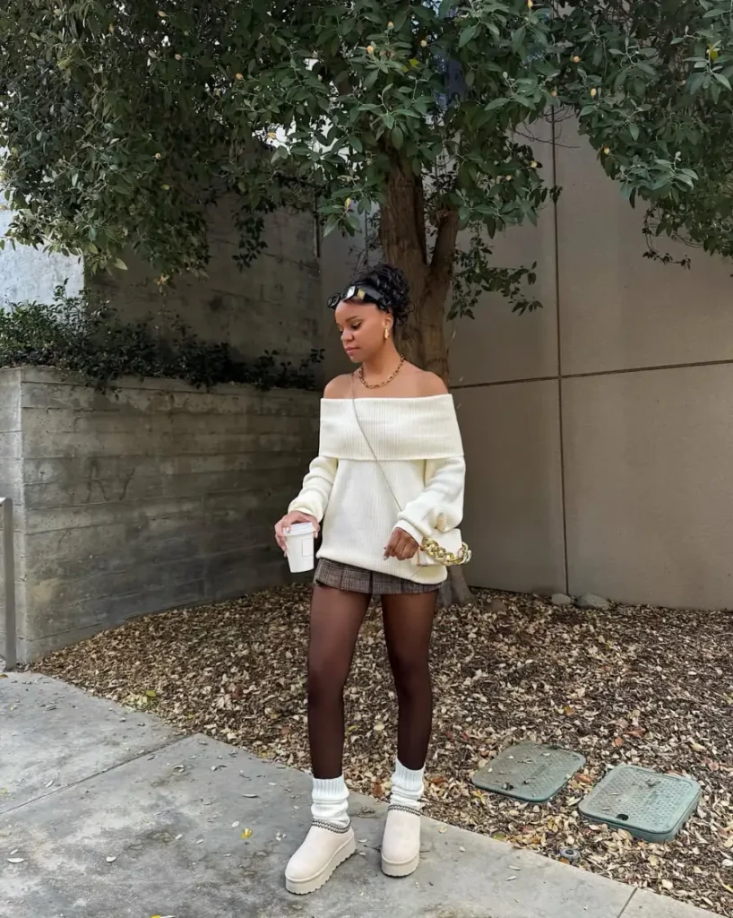 Black woman wearing an oversized cream off-shoulder sweater with a plaid mini skirt, sheer tights, cream platform boots, scrunched socks, and gold accessories for a cozy fall look.