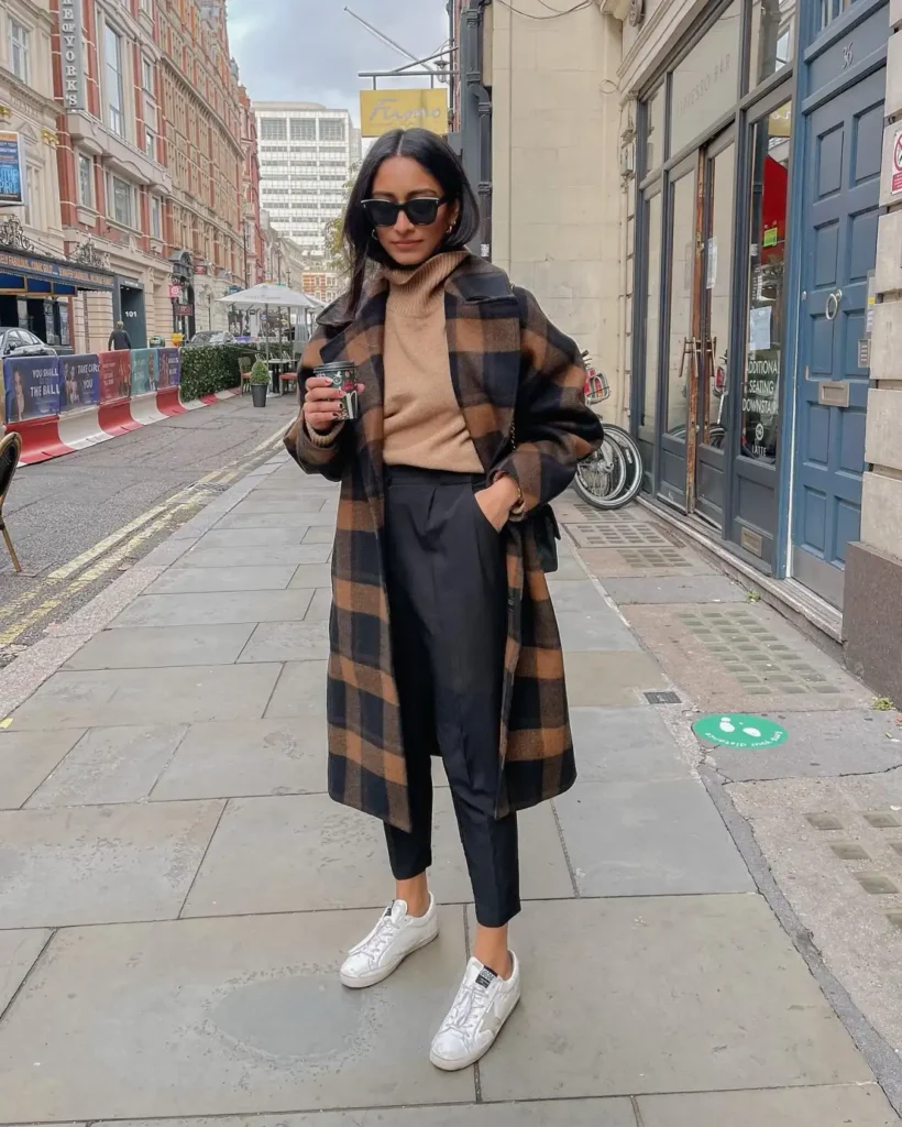Woman wearing a long chocolate-brown and black plaid coat over a camel turtleneck sweater, tailored black ankle trousers, and white sneakers for a casual chic winter street-style look.