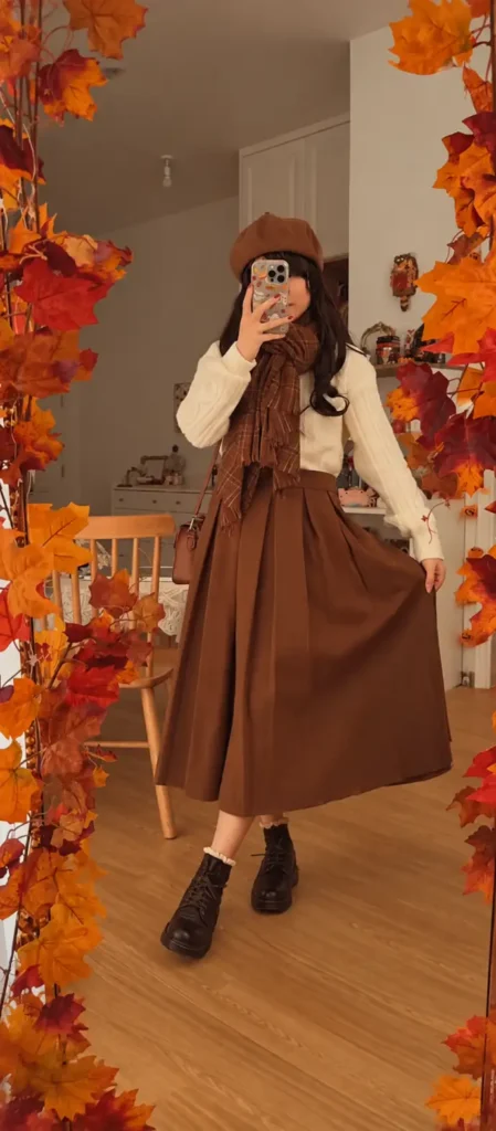 Cream cable-knit sweater styled with a full brown pleated maxi skirt, plaid scarf, brown beret, and dark lace-up boots.