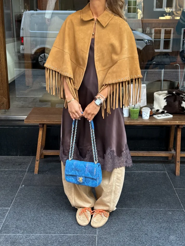 Woman wearing a tan fringe cape over a brown lace-trim slip dress layered with light wide-leg jeans and a bright blue bag.