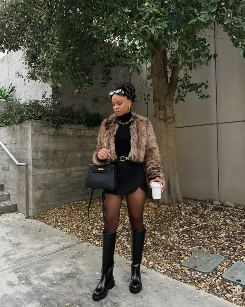 Black woman wearing a cropped brown faux-fur jacket over a fitted black turtleneck and black mini skirt with sheer tights, knee-high black boots, and a structured handbag.