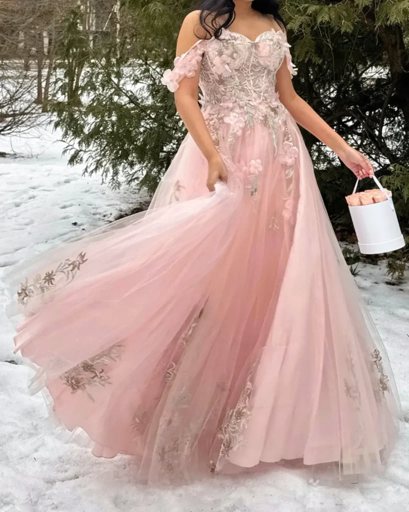 Woman wearing a blush pink off-shoulder tulle gown with delicate floral embroidery, a fitted corset bodice, and a flowing skirt in a snowy winter setting.