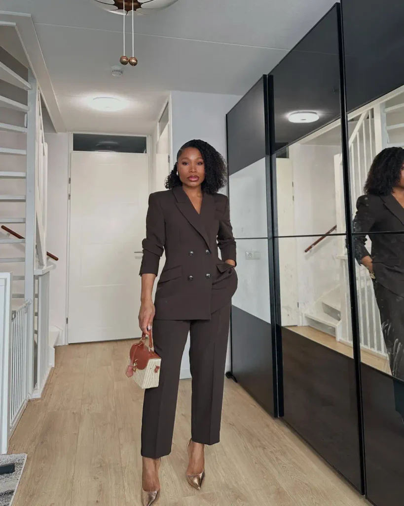 Black woman wearing a chocolate brown double-breasted blazer with matching cropped tailored trousers, metallic pointed heels, and a small structured handbag for professional corporate style.