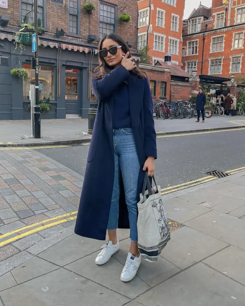 Woman styling a long navy wool coat over a matching dark knit sweater, cropped blue jeans, white sneakers, and a canvas tote bag for an effortless everyday winter outfit.