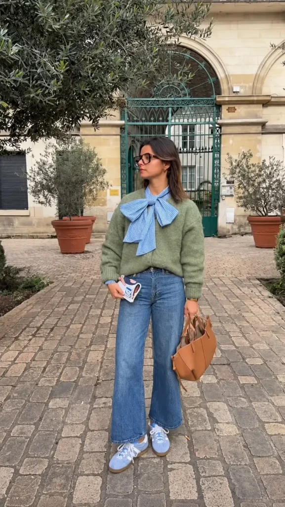 Petite woman wearing a sage green sweater layered over a blue gingham bow blouse with wide-leg jeans, blue sneakers, and a tan handbag.
