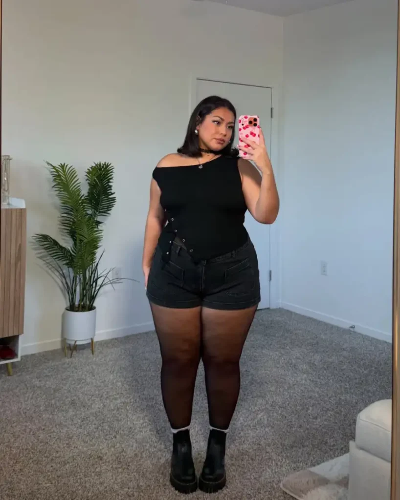 Plus size woman wearing a black off-shoulder top with black denim shorts, sheer tights, and chunky black ankle boots for an edgy fall outfit.