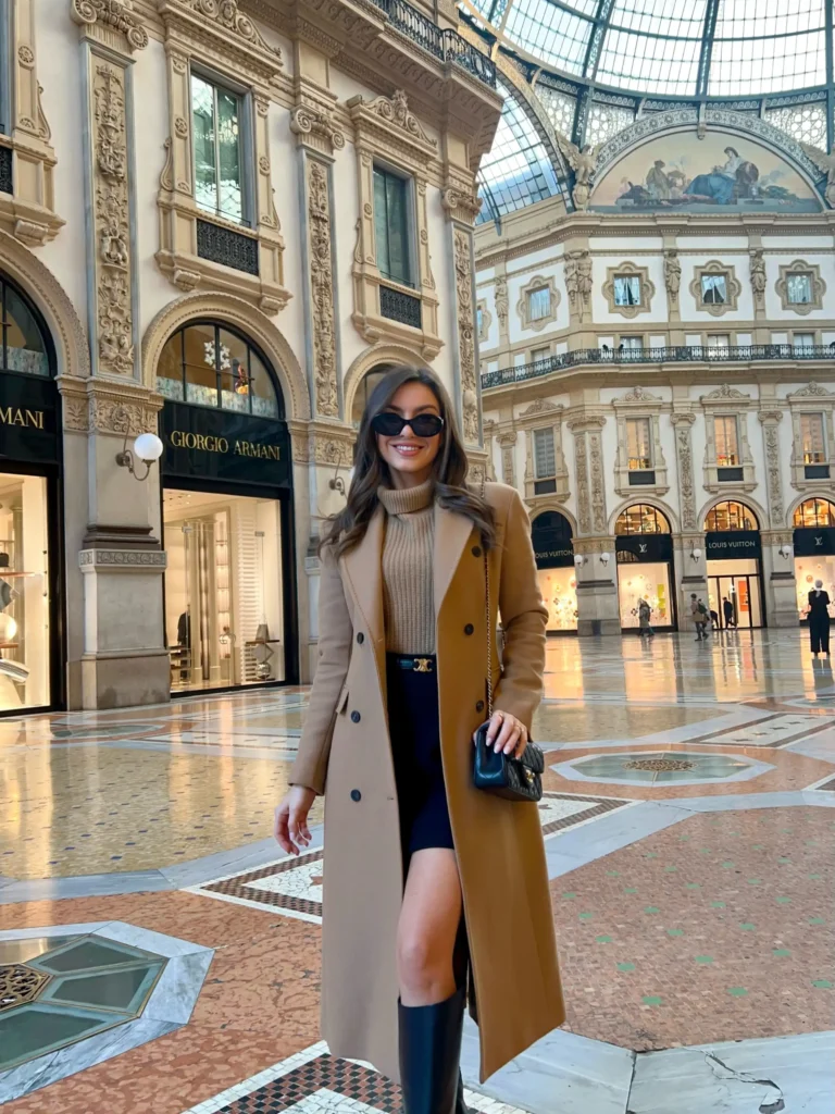 Long camel coat layered over a beige turtleneck and black mini skirt with knee-high boots for an elegant fall outfit for women.