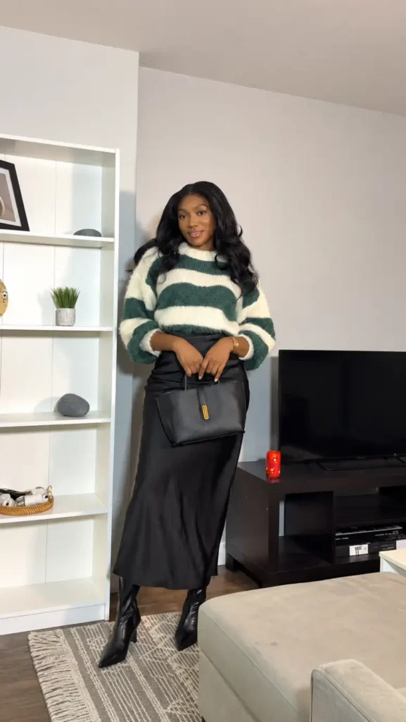 Black woman wearing a fuzzy green and cream striped sweater with a black satin maxi skirt, pointed black boots, and a structured black handbag for a stylish fall outfit.