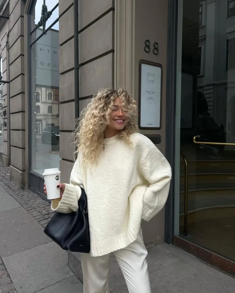 Woman wearing an oversized ivory chunky-knit sweater with relaxed cream trousers and a slouchy black leather handbag for a cozy monochrome winter outfit.