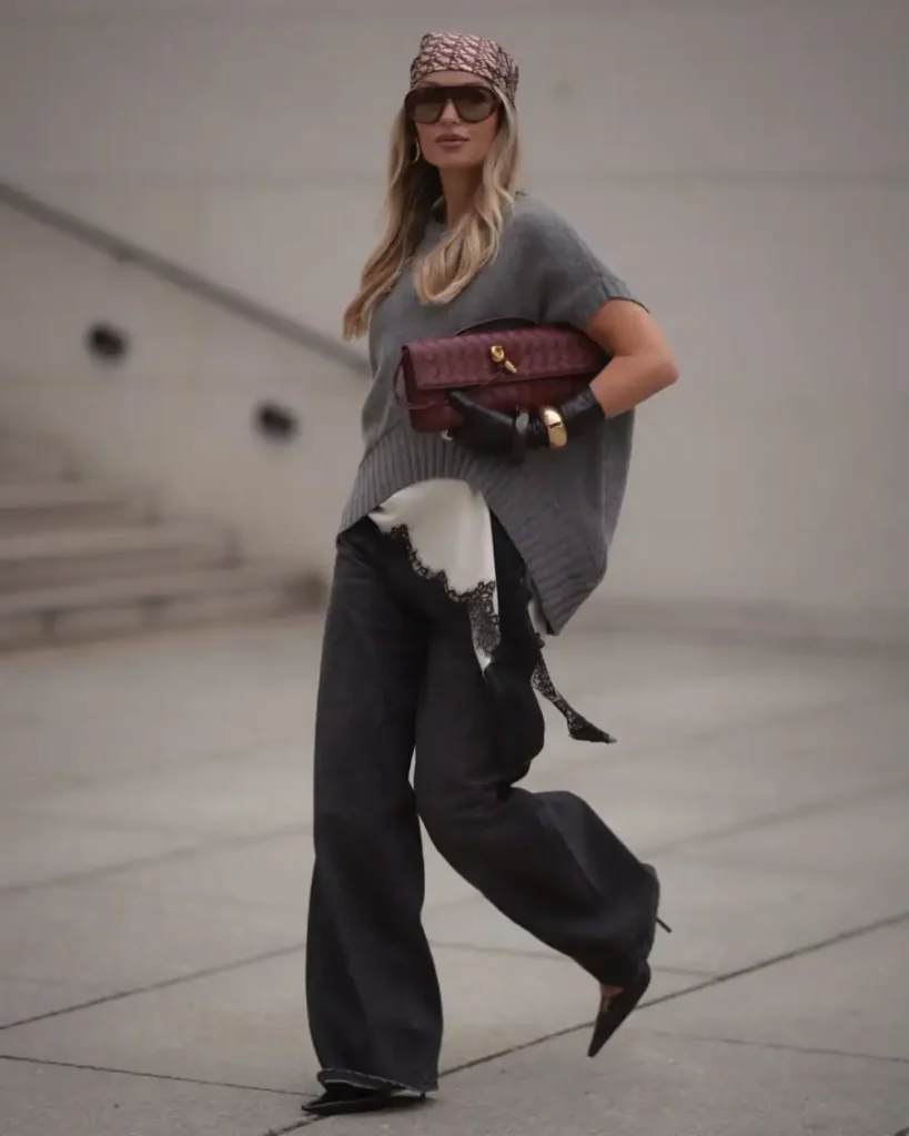 Woman wearing an oversized gray knit top with dark wide-leg jeans, a burgundy woven clutch, patterned headscarf, black gloves, and pointed shoes for a fashion-forward fall look.