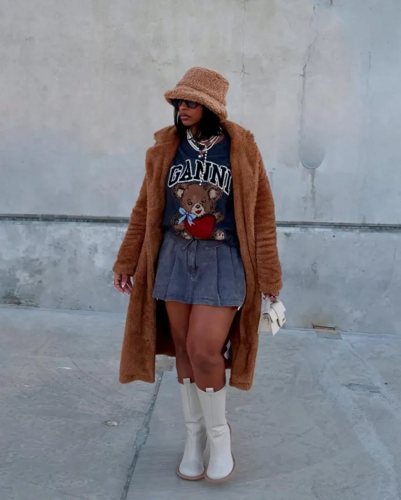 Black woman wearing a long brown teddy coat over a graphic sweater and gray mini skirt with cream knee-high boots, layered necklaces, sunglasses, and a fuzzy bucket hat.