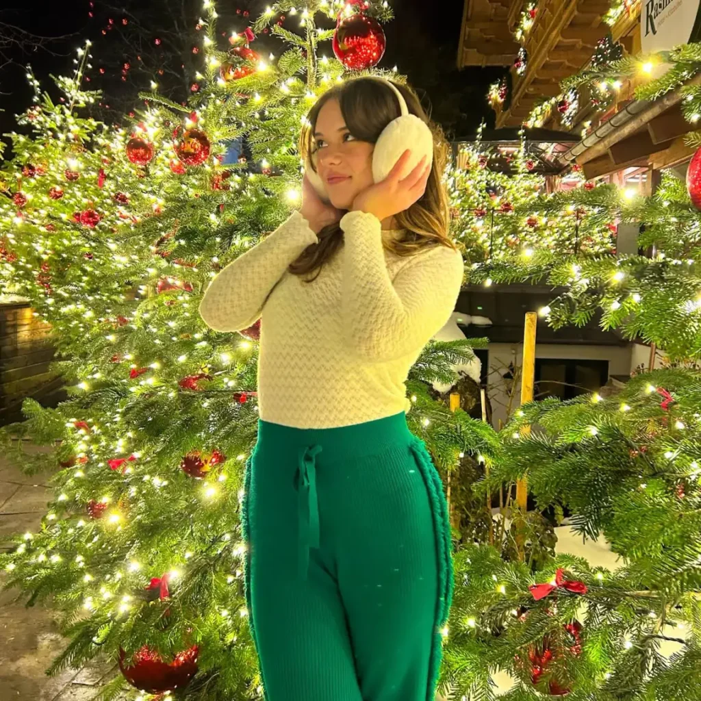 Woman wearing a fitted cream knit top with emerald-green ribbed drawstring trousers and fluffy white earmuffs while standing among illuminated Christmas trees for a cozy holiday outfit.