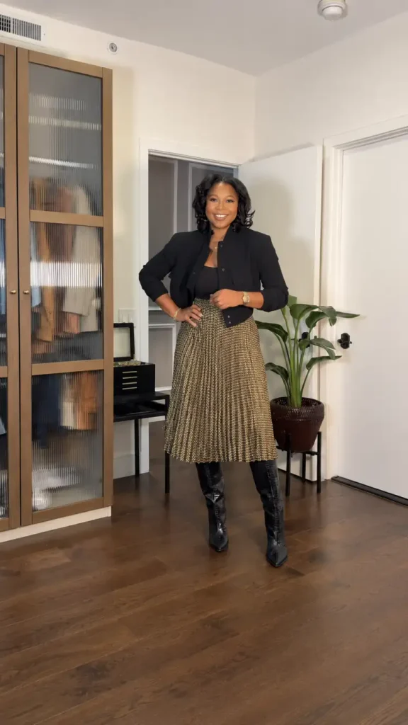 Black woman wearing a cropped black jacket over a fitted black top with a gold patterned pleated midi skirt and tall black boots for a classy fall office outfit.