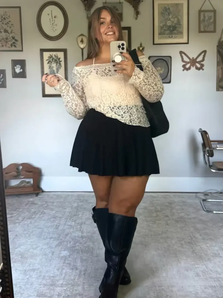 Plus size woman wearing a cream off-shoulder lace top with a black mini skirt, tall black boots, and a black tote bag for a feminine fall look.