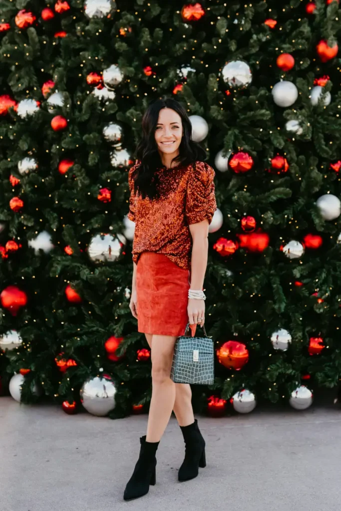 Woman wearing a rust orange sequin puff-sleeve top with a terracotta mini skirt, black ankle boots, and a structured gray handbag for a festive Christmas outfit.