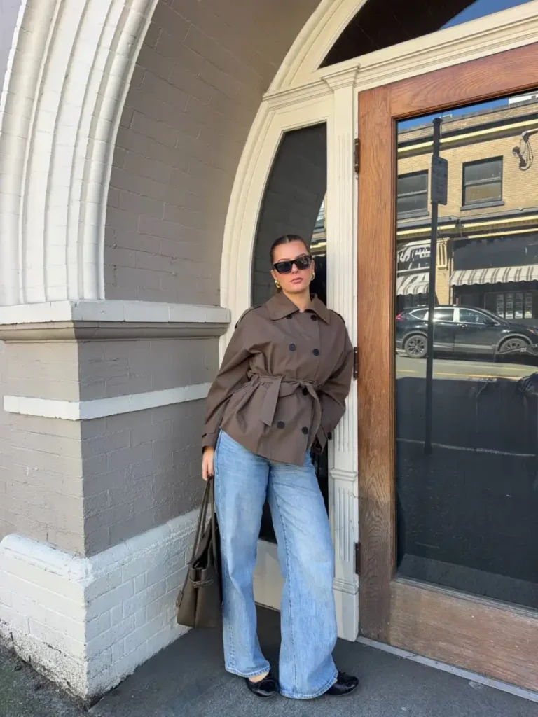 Woman wearing a short brown belted trench coat with light-wash wide-leg jeans, black ballet flats, sunglasses, and a dark tote for a casual fall outfit.