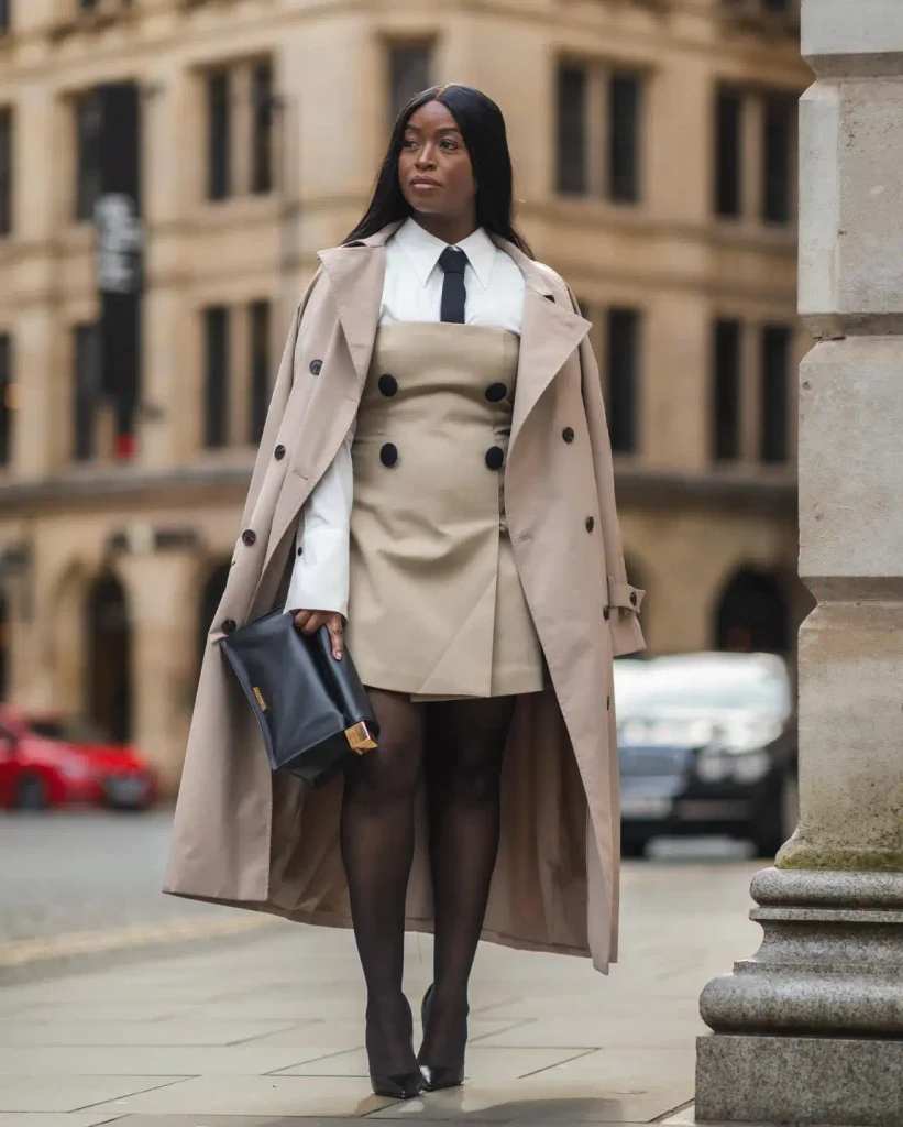 Black woman wearing a beige double-breasted mini dress over a white collared shirt and black tie with a long trench coat, sheer tights, and pointed black heels.