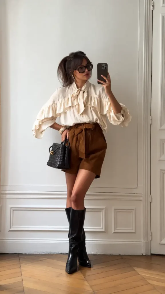 Petite woman wearing an ivory ruffled tie-neck blouse with high-waisted brown suede shorts, black knee-high boots, and a woven black handbag.
