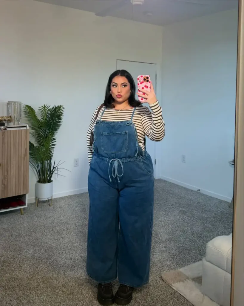 Plus size woman wearing wide-leg denim overalls over a cream and brown striped long-sleeve top with dark chunky shoes for a casual fall outfit.