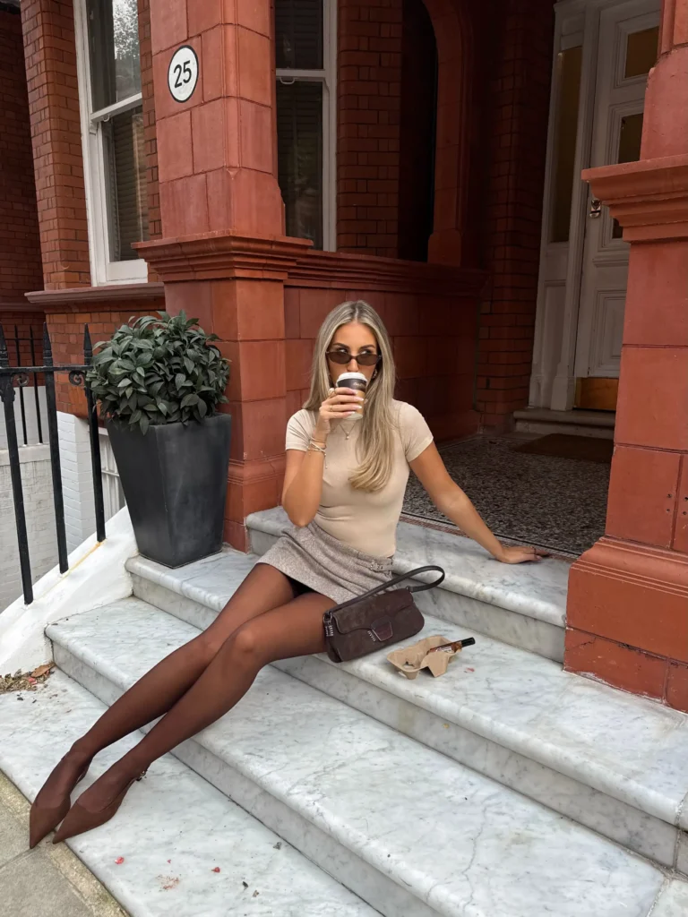 Beige fitted knit and neutral mini skirt styled with sheer brown tights, pointed brown shoes, and a suede handbag for a fall outfit.