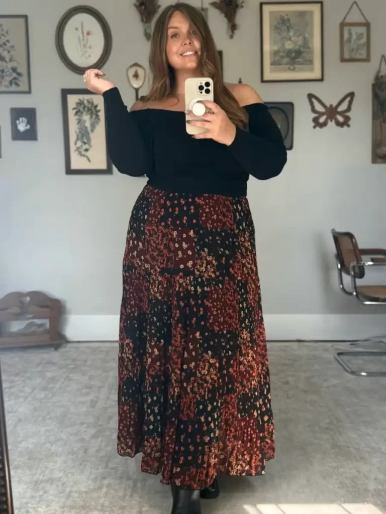 Plus size woman wearing a black off-shoulder top with a dark floral maxi skirt featuring rust and burgundy tones and black ankle boots for fall.
