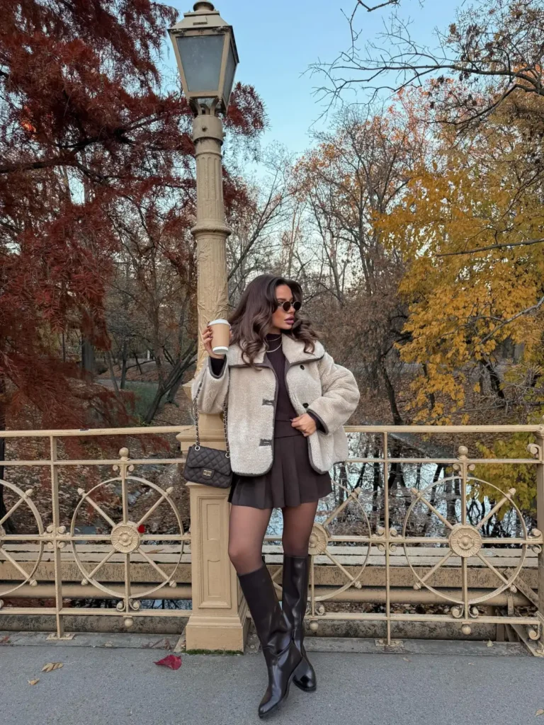 Cream shearling jacket layered over a brown turtleneck and pleated mini skirt with sheer tights and knee-high boots for a cozy fall outfit.