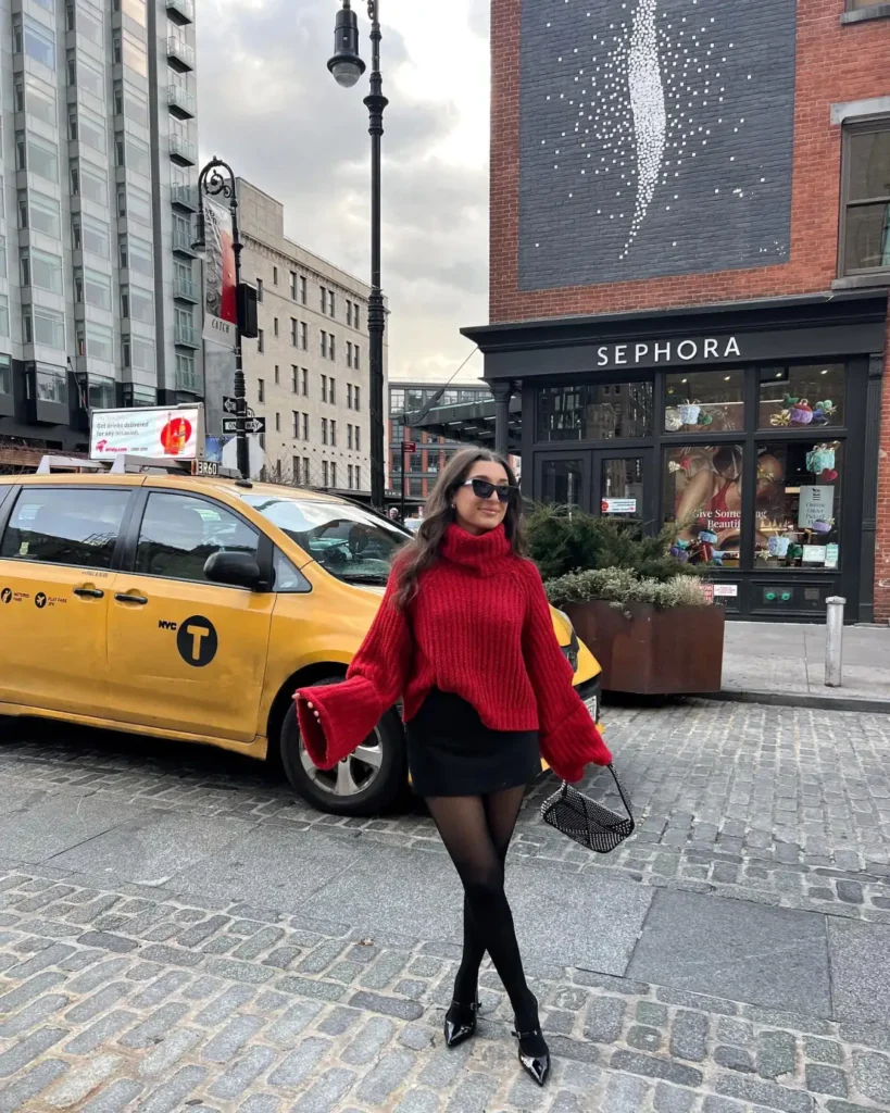 Woman styling an oversized cherry-red chunky turtleneck sweater with dramatic sleeves, a black mini skirt, sheer black tights, pointed heels, and a small handbag for a dressy winter outfit.