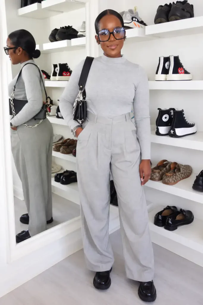 Black woman wearing a fitted light-gray mock-neck top with matching high-waisted pleated wide-leg trousers, chunky black loafers, oversized glasses, and a black shoulder bag.
