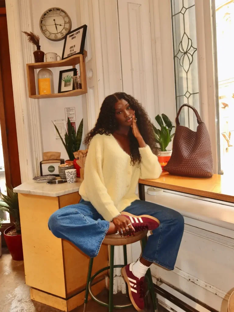 Black woman wearing a fluffy cream cardigan with relaxed blue jeans, burgundy sneakers, white socks, and a woven brown tote for a casual fall outfit.