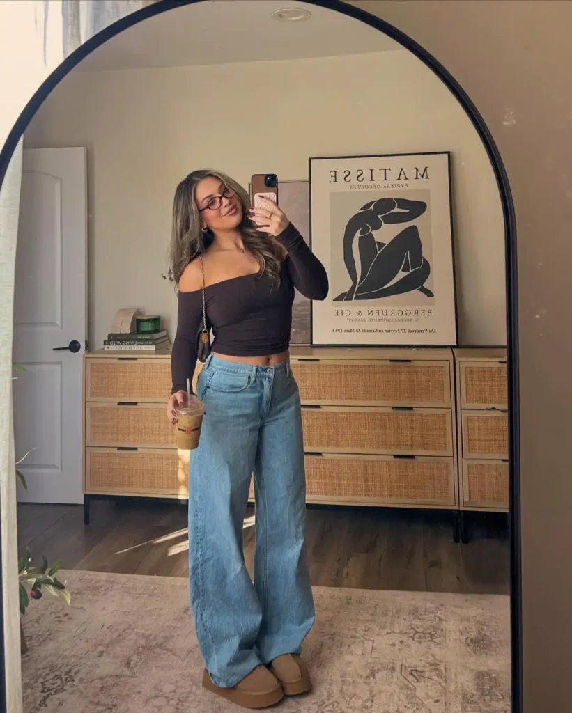 Woman wearing a chocolate brown off-shoulder top with light-wash baggy jeans, tan platform slip-on shoes, and a small shoulder bag for a casual Friendsgiving outfit.