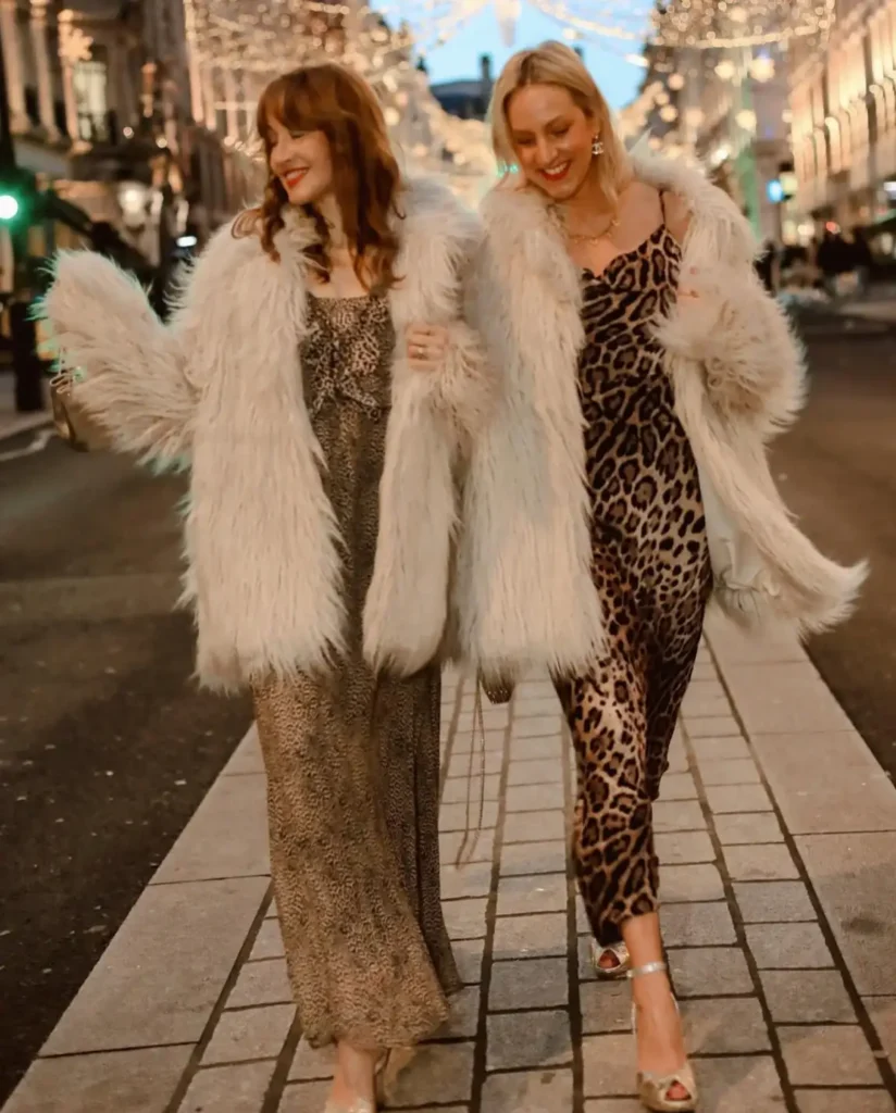 Two women wearing leopard print midi dresses with fluffy ivory faux fur coats and metallic heels for glamorous winter Christmas party outfits.