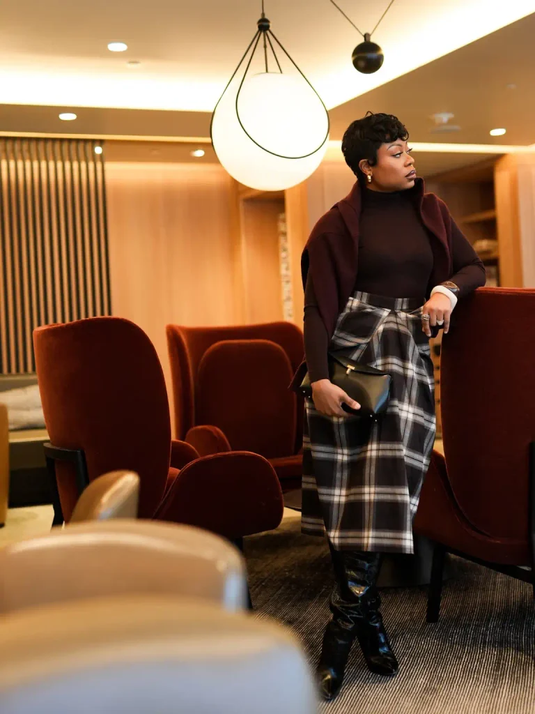 Black woman styling a plaid midi skirt with a burgundy turtleneck, matching shoulder-draped layer, black knee-high boots, and a clutch for an elegant fall office look.