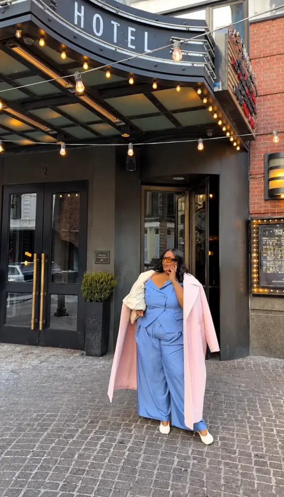 Plus size Black woman wearing a powder blue vest and wide-leg trousers with a long blush pink coat for a polished fall outfit.