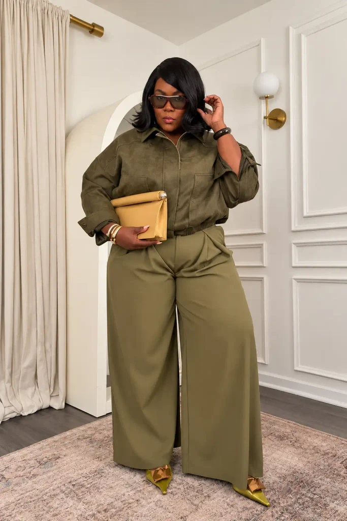 Plus size Black woman styling olive green wide-leg trousers with a matching suede-look jacket, mustard clutch, and pointed heels for a sophisticated fall outfit.