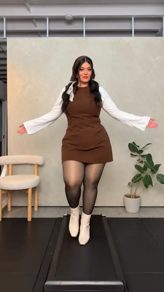 Mid size woman wearing a chocolate brown mini dress over a white flared-sleeve top with sheer tights, cream ankle boots, and a brown headscarf.