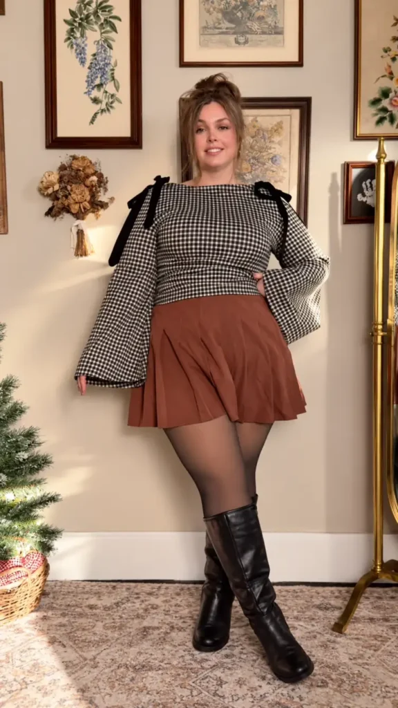 Plus size woman wearing a black and cream gingham top with statement sleeves, rust brown pleated mini skirt, sheer tights, and tall black boots.