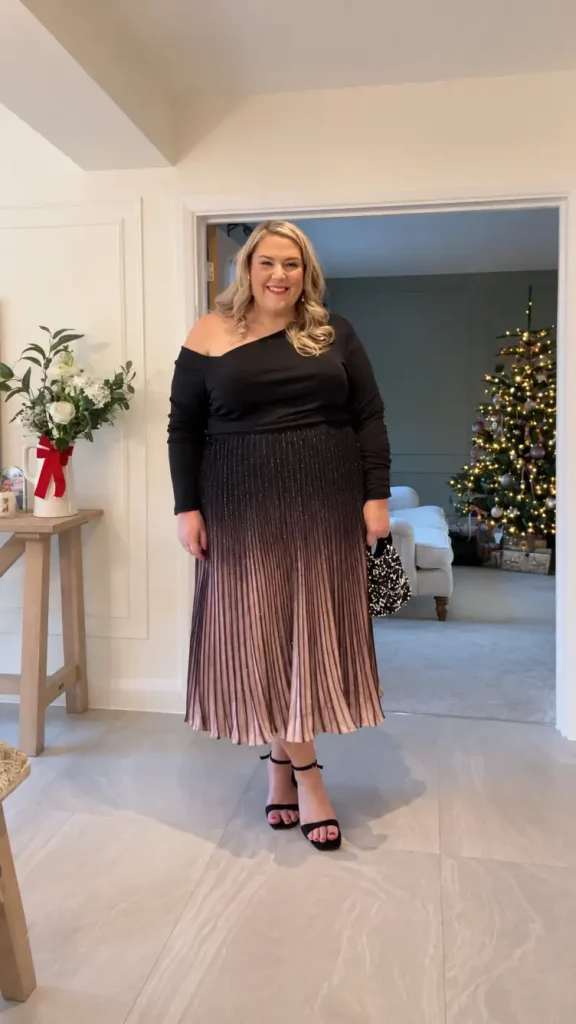 Plus-size woman wearing a black one-shoulder top with a black and rose gold ombre pleated midi skirt, black heels, and a sparkly handbag for a classy Christmas look.