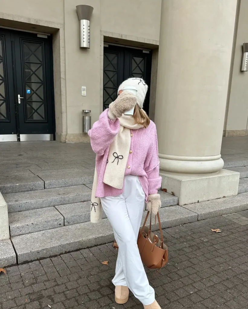 School girl wearing an oversized pink cardigan with white wide-leg trousers, a cream bow-patterned scarf, white beanie, fluffy mittens, and beige platform shoes.