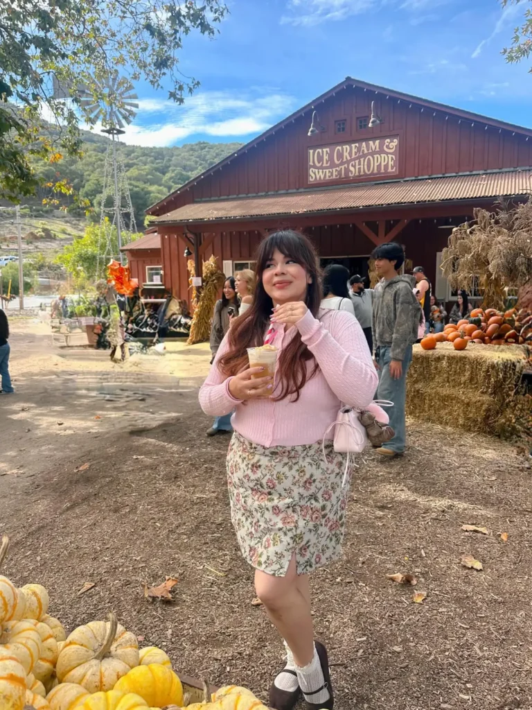Plus size fall outfit with a pink cardigan, cream floral skirt, white socks, brown Mary Jane shoes, and a small pink shoulder bag at a pumpkin patch.