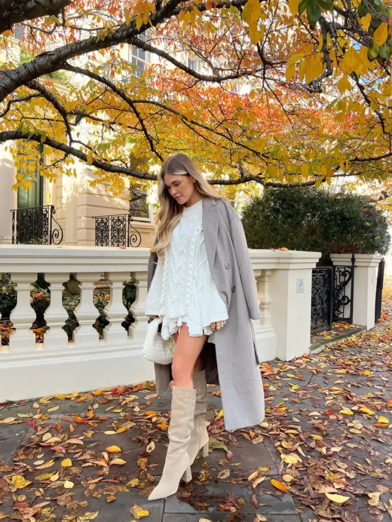 White cable knit sweater layered under a long gray coat with light knee-high boots and a neutral handbag for cozy fall outfits for women.