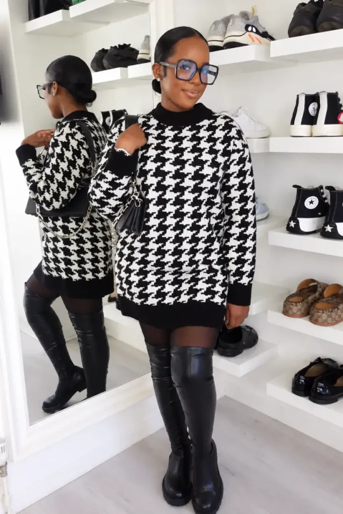 Black woman wearing an oversized black-and-white houndstooth sweater dress with sheer black tights, over-the-knee black boots, glasses, and a black shoulder bag.
