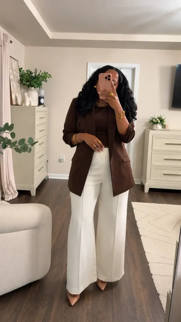 Black woman wearing a chocolate brown blazer over a matching fitted top with cream wide-leg trousers, nude pointed heels, and gold accessories for a classy fall office outfit.