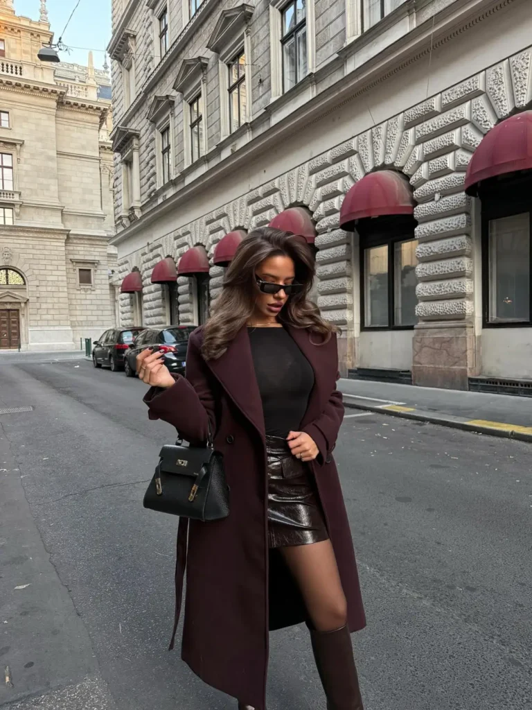Woman wearing a deep burgundy long coat, black top, dark chocolate-brown leather mini skirt and brown knee-high boots for a chic fall outfit.