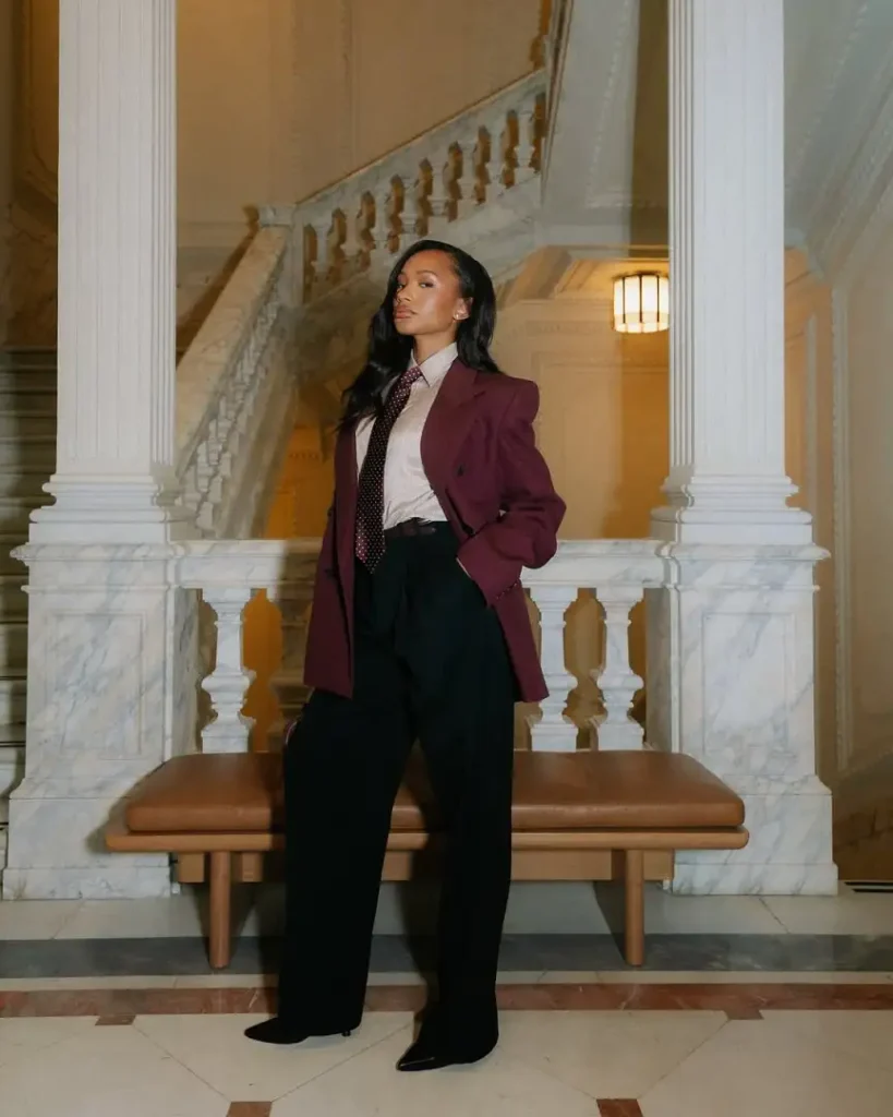 Black woman wearing a burgundy tailored blazer with a striped button-down shirt, patterned necktie, black wide-leg trousers, and pointed shoes for a powerful corporate outfit.