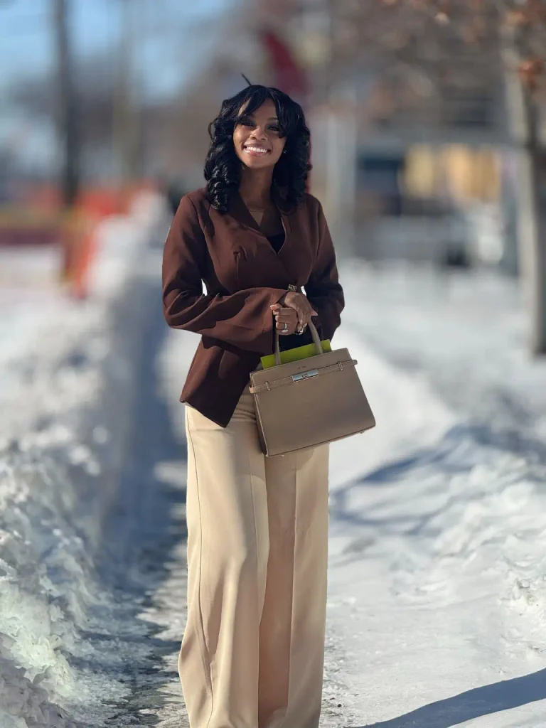 Black woman wearing a structured chocolate brown blazer with a defined waist, cream tailored trousers, and a taupe top-handle handbag for elegant business professional fashion.