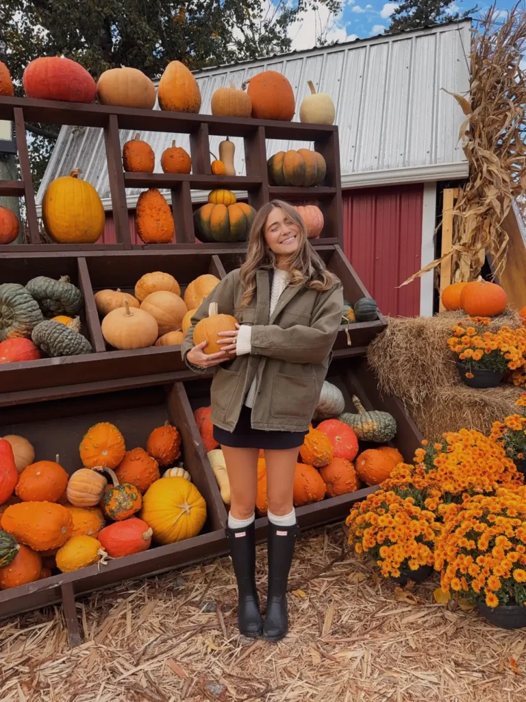 Woman wearing a muted olive-green utility jacket, cream cable-knit sweater, black mini skirt and black knee-high rain boots at a pumpkin patch.
