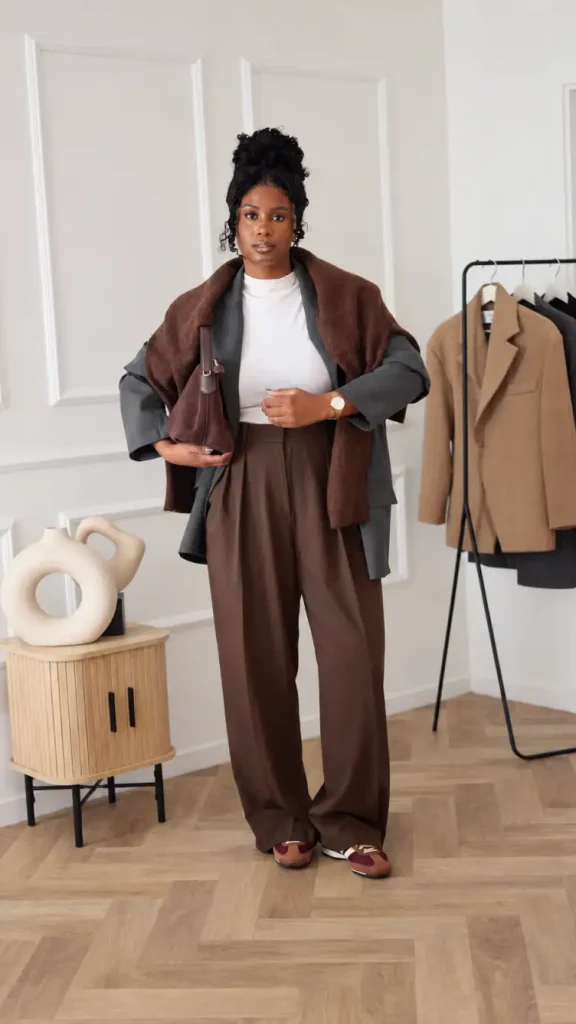 Black woman wearing a charcoal blazer over a white top with chocolate brown wide-leg trousers, a brown sweater draped over her shoulders, and burgundy sneakers.