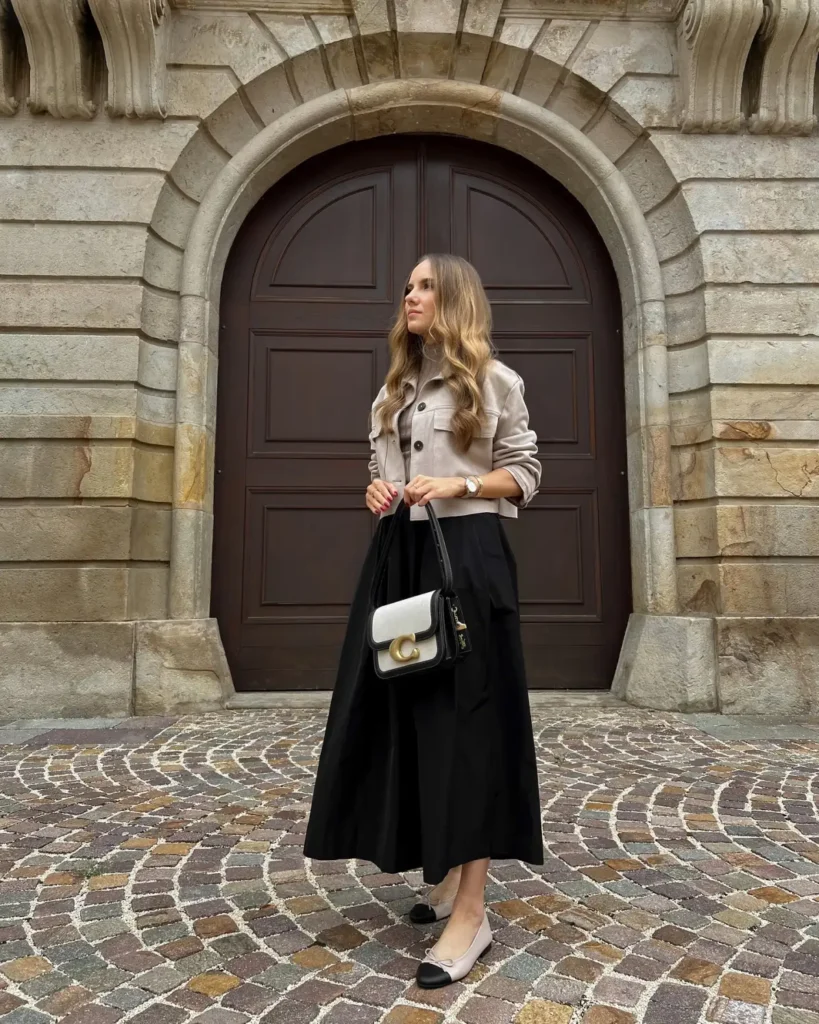 Woman wearing a light taupe-beige cropped jacket, black midi skirt and beige ballet flats with black cap toes for an elegant neutral fall outfit.