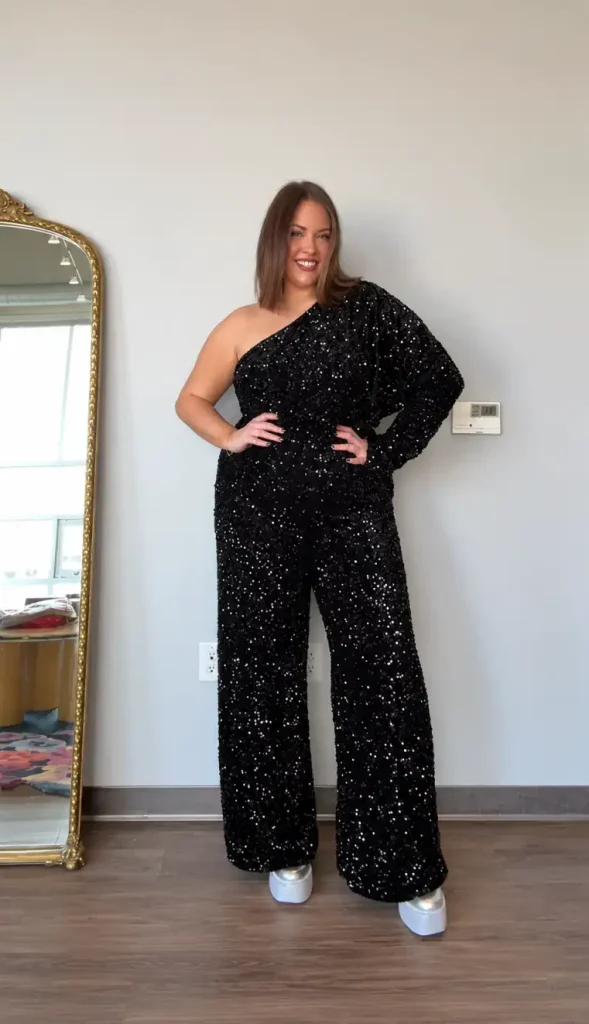 Woman wearing a black sequin wide-leg jumpsuit with an asymmetric one-shoulder neckline, one long sleeve, and silver platform shoes for a glamorous New Year's Eve celebration.